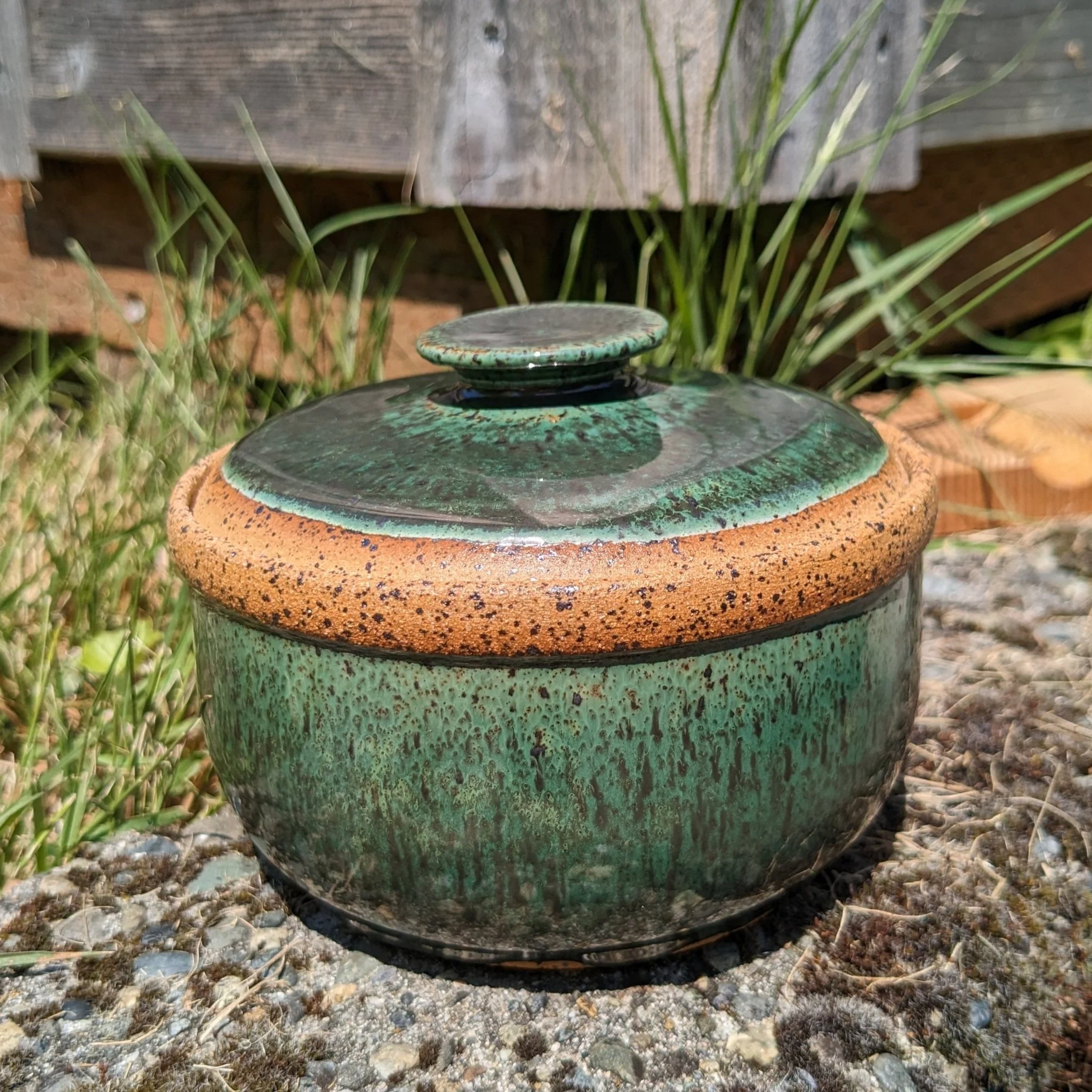 A small, round ceramic container with a lid, glazed in green and brown with a speckled texture, placed outdoors on a gravel surface.