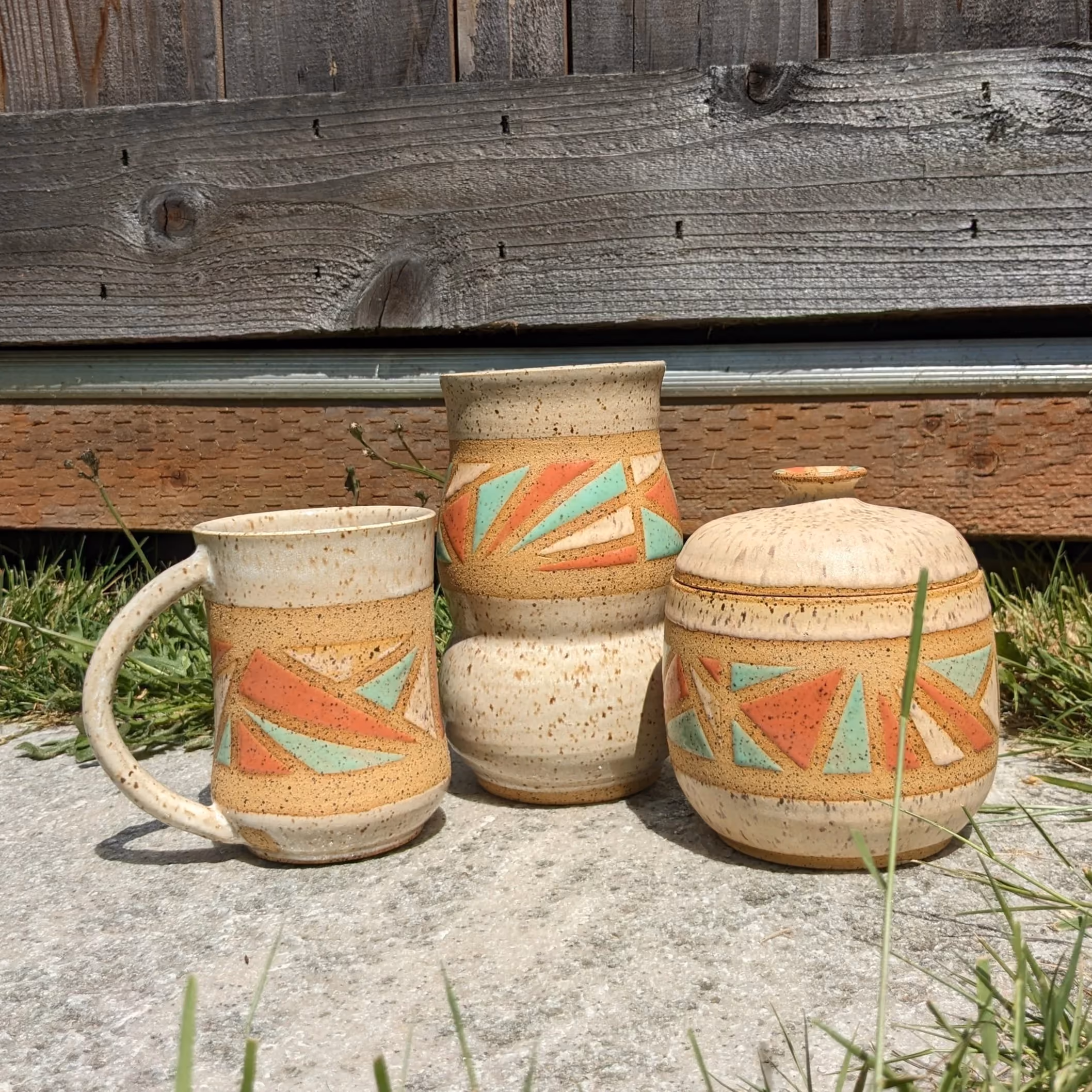 Three handcrafted ceramic pottery pieces with geometric designs in pastel colors, placed outdoors on the ground in front of a wooden fence.