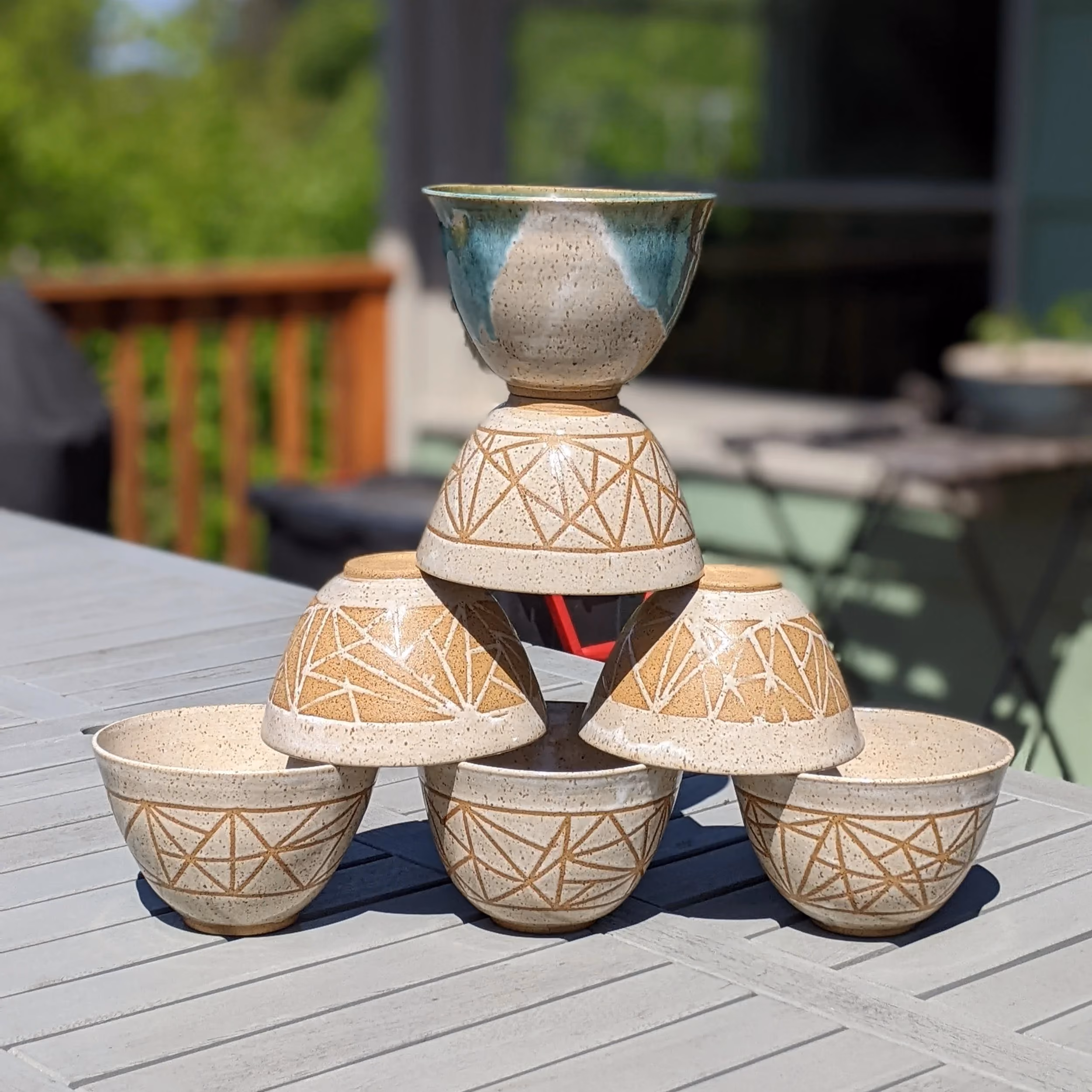 Ceramic bowls stacked in a pyramid on a wooden outdoor table, with a background of a deck and greenery.