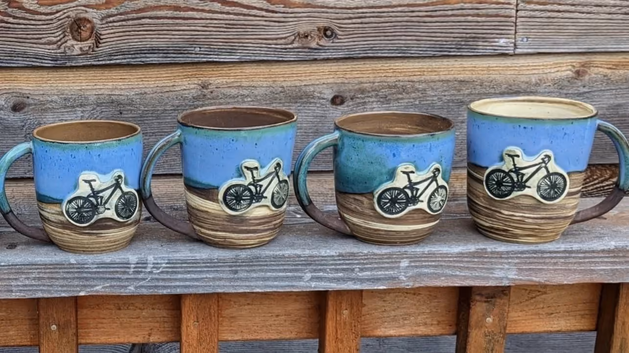 Four ceramic coffee mugs with bicycle decorations, placed on a weathered wooden shelf against a wooden wall.
