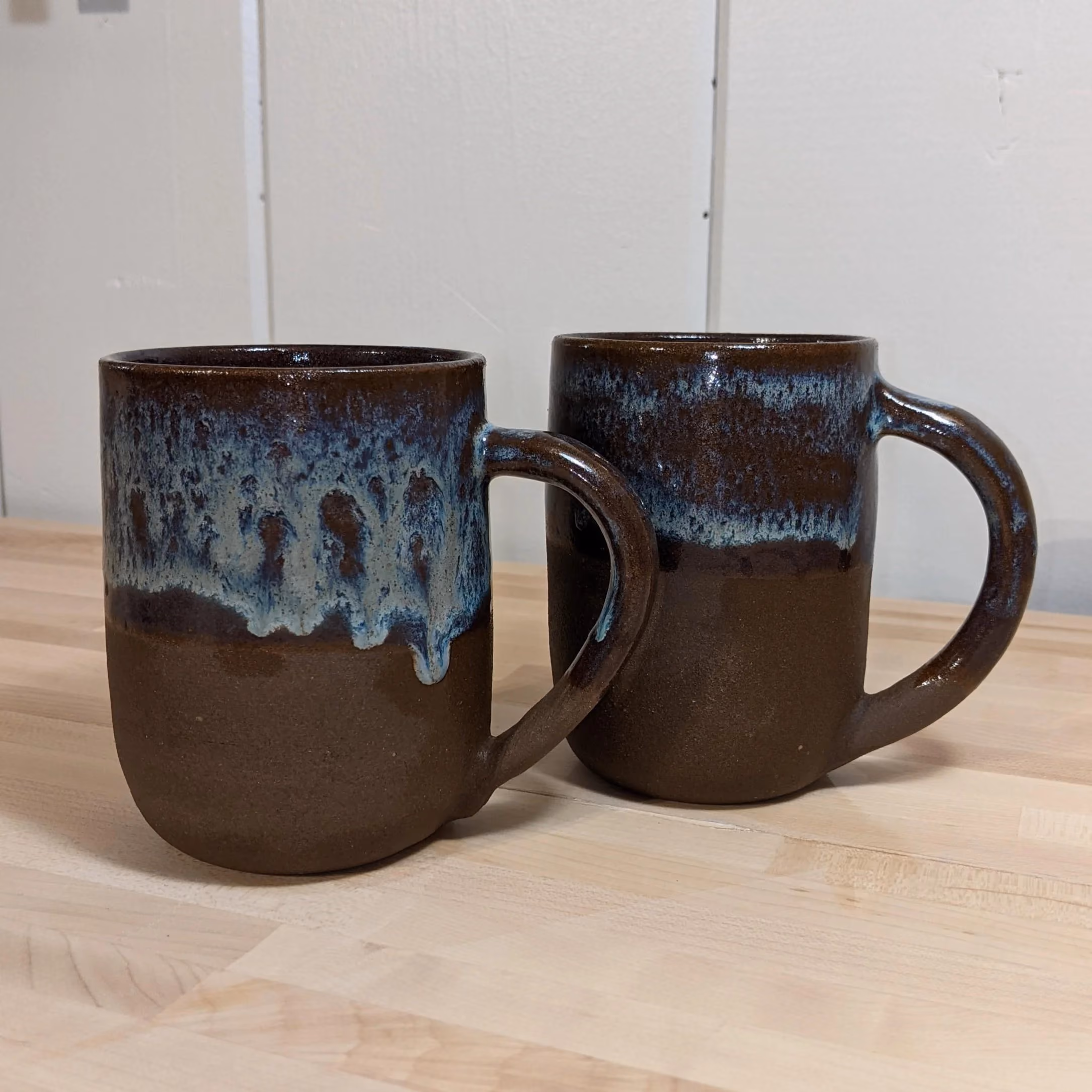 Two handcrafted ceramic coffee mugs with brown and blue glaze, placed on a light wooden surface against a white wall.