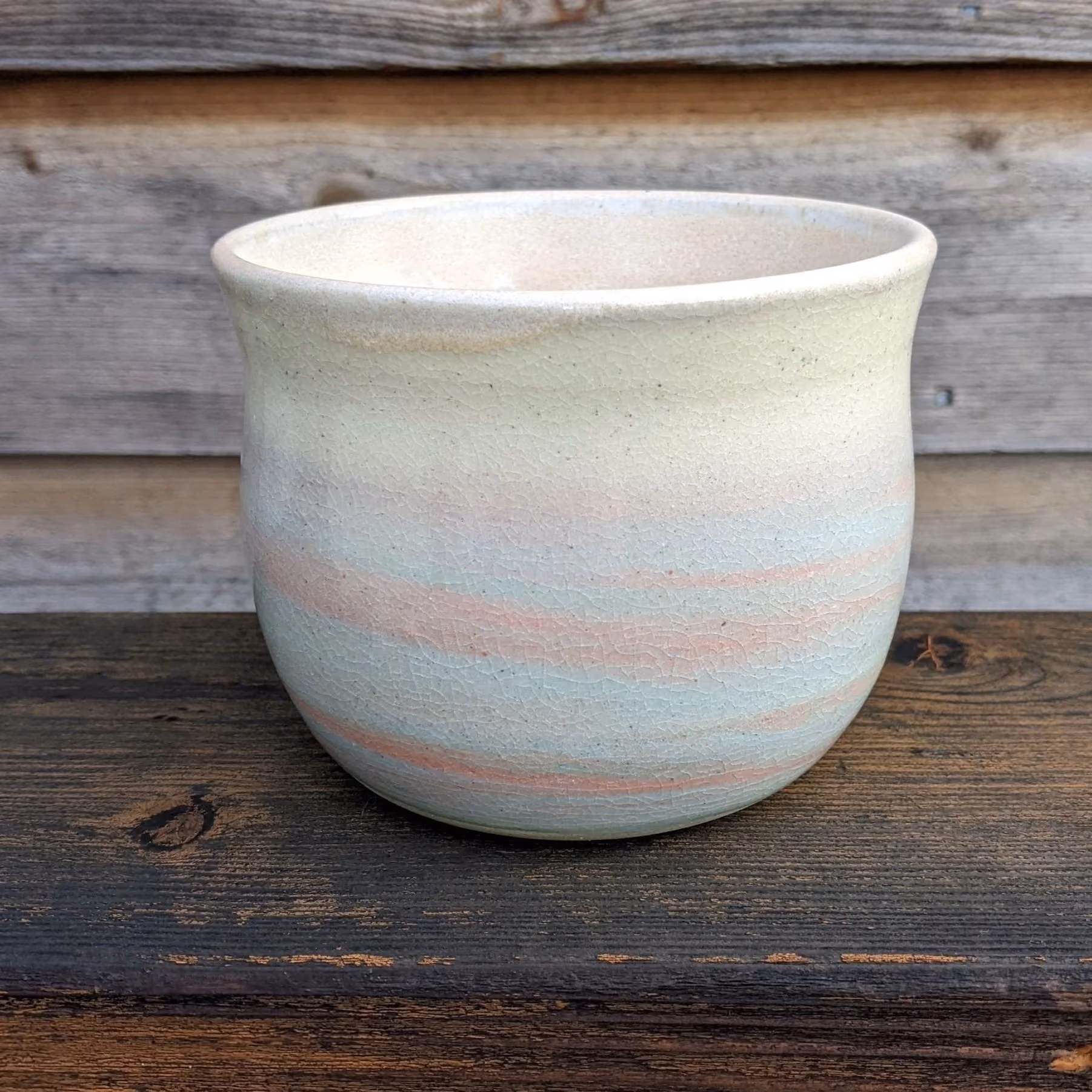 A pastel-colored ceramic flowerpot with a crackled finish, placed on a dark wooden surface against a weathered wooden wall background.