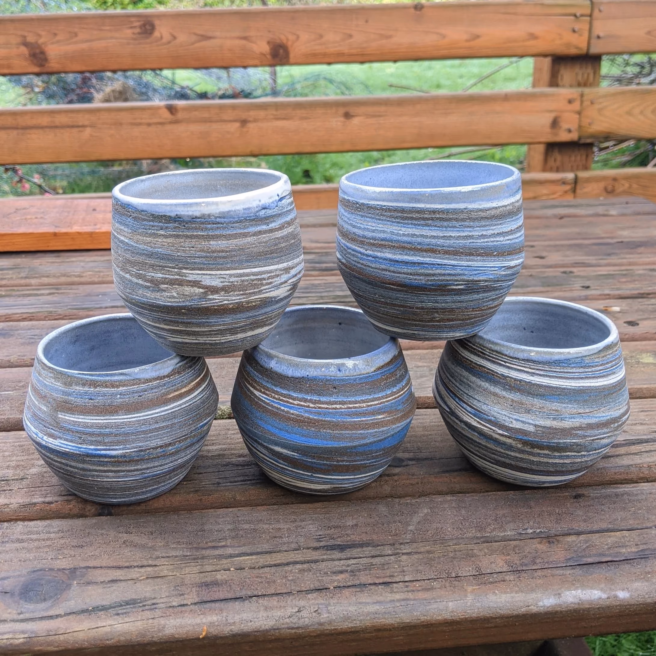Five marbled ceramic cups arranged on a wooden outdoor table with a wooden fence and greenery in the background.