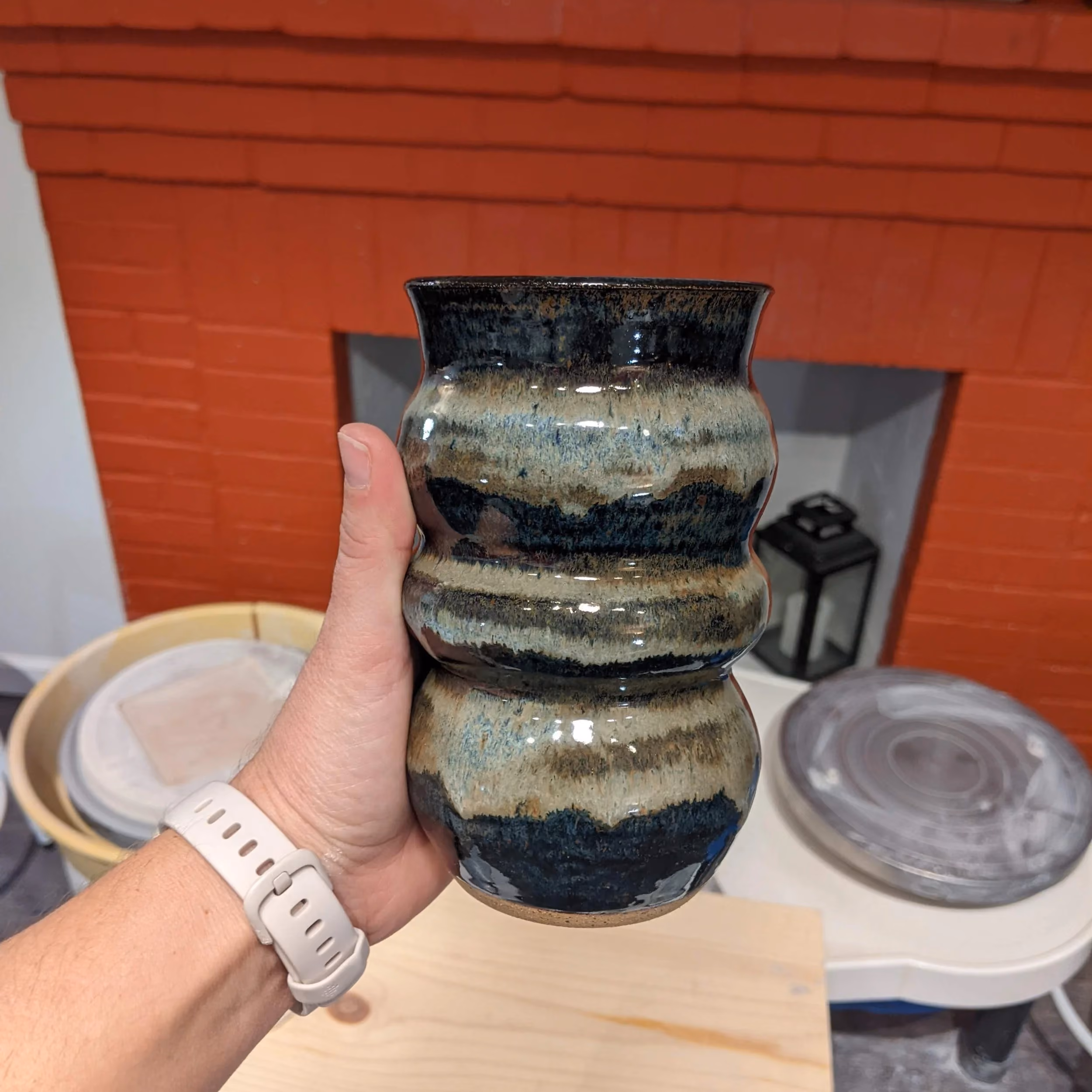 A hand holding a ceramic vase with a wavy, textured shape and a glaze in black, beige, and blue tones. In the background, there is a table with decorative objects, including a lid and a lantern, and a red brick wall.