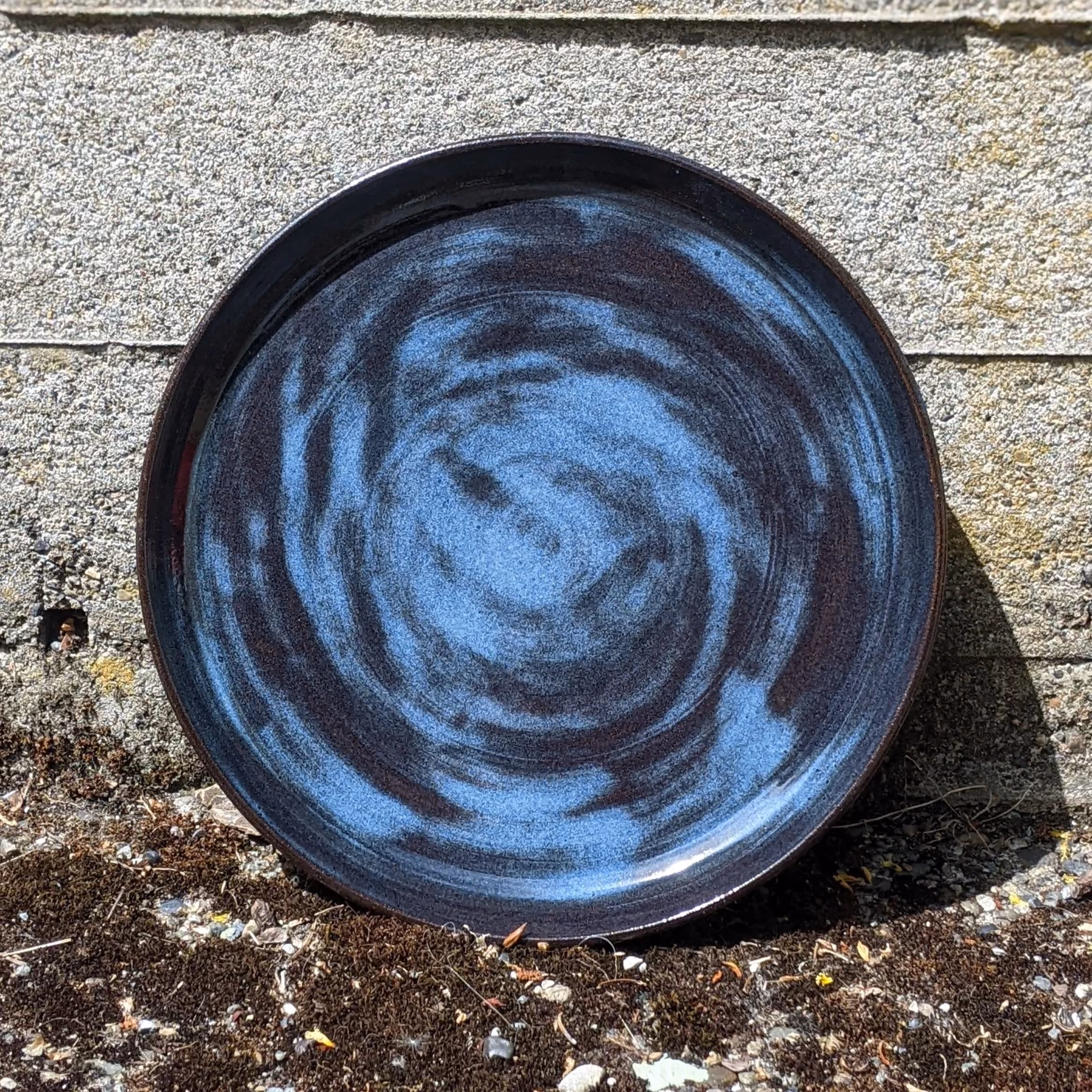A round ceramic dish with a dark and blue swirling glaze pattern, resting on a concrete surface.
