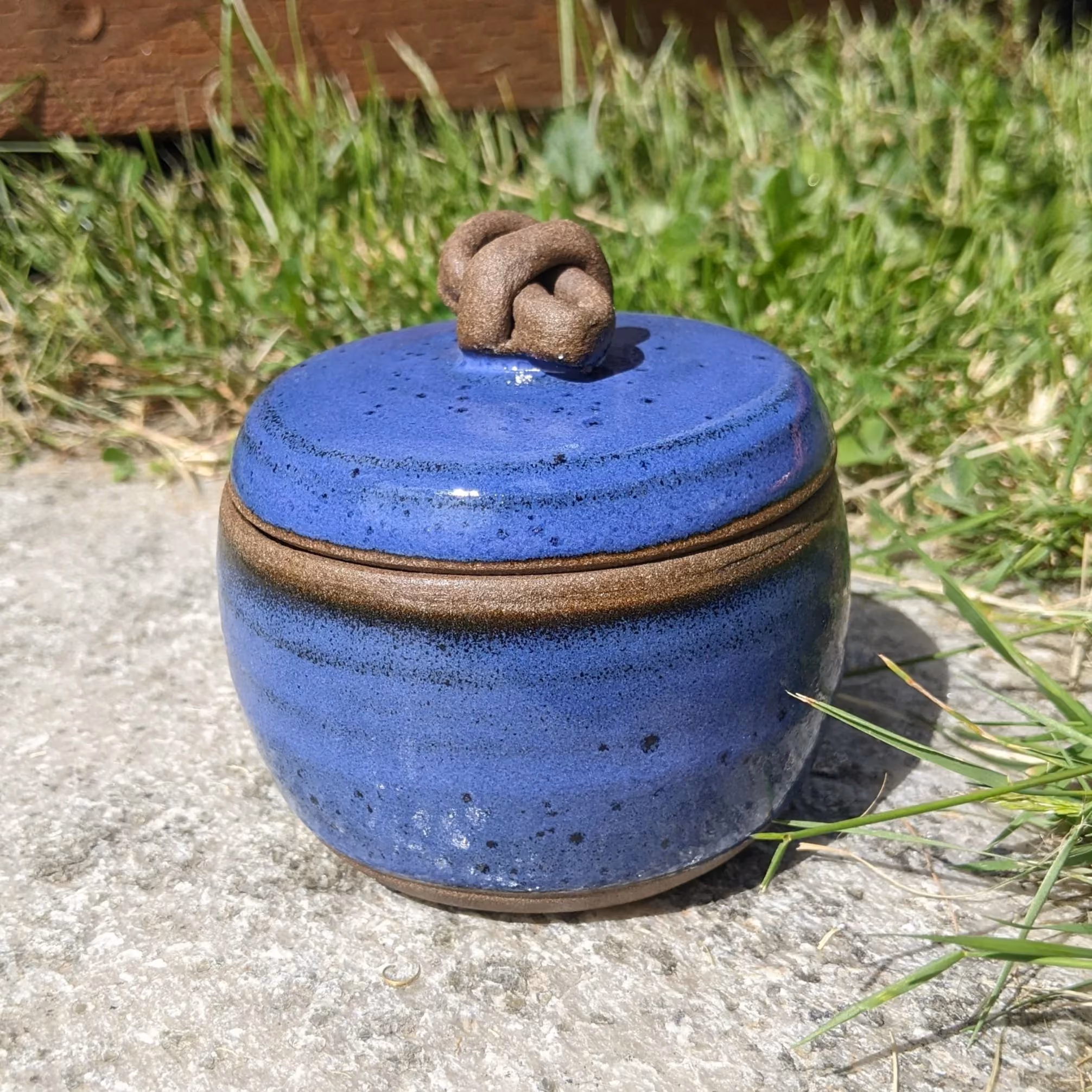 A small blue ceramic jar with a matching lid, decorated with a clay knot on top, sitting outdoors on a concrete surface surrounded by grass.