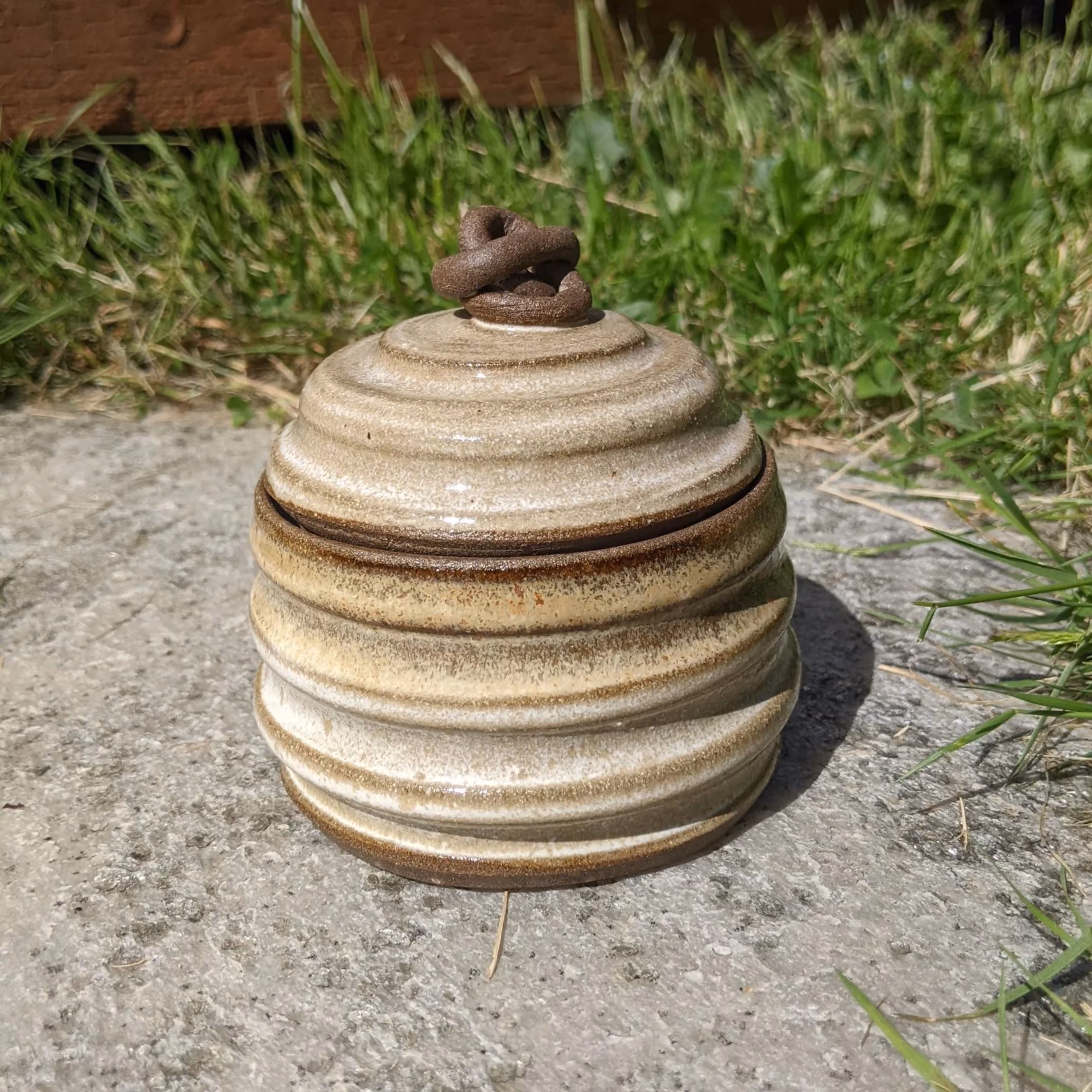 A ceramic lid with a small, twisted brown handle on top, placed outdoors on a concrete surface next to green grass.