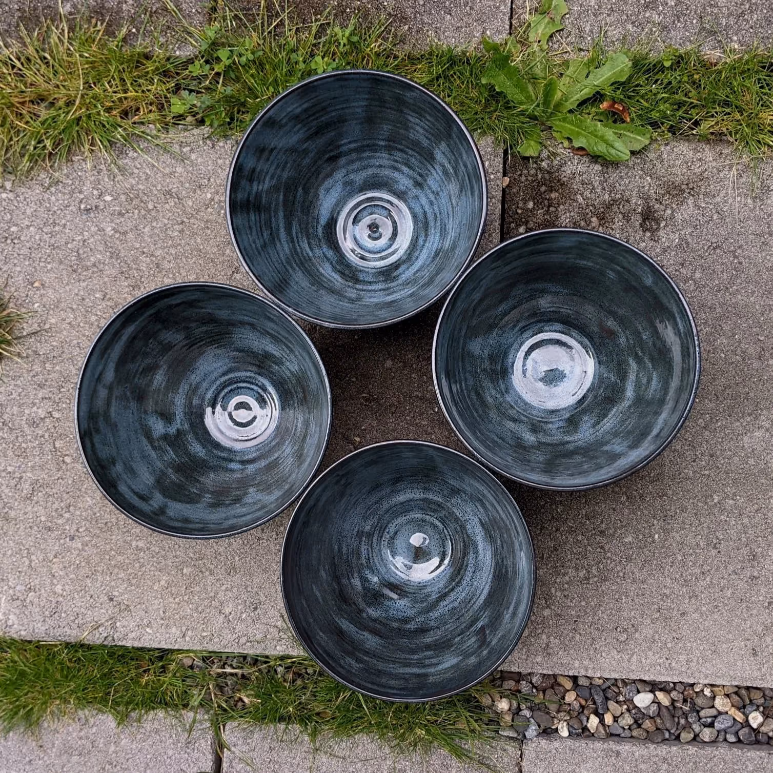 Four large black ceramic bowls with a swirl pattern inside, placed on a concrete surface outdoors with grass and small plants nearby.