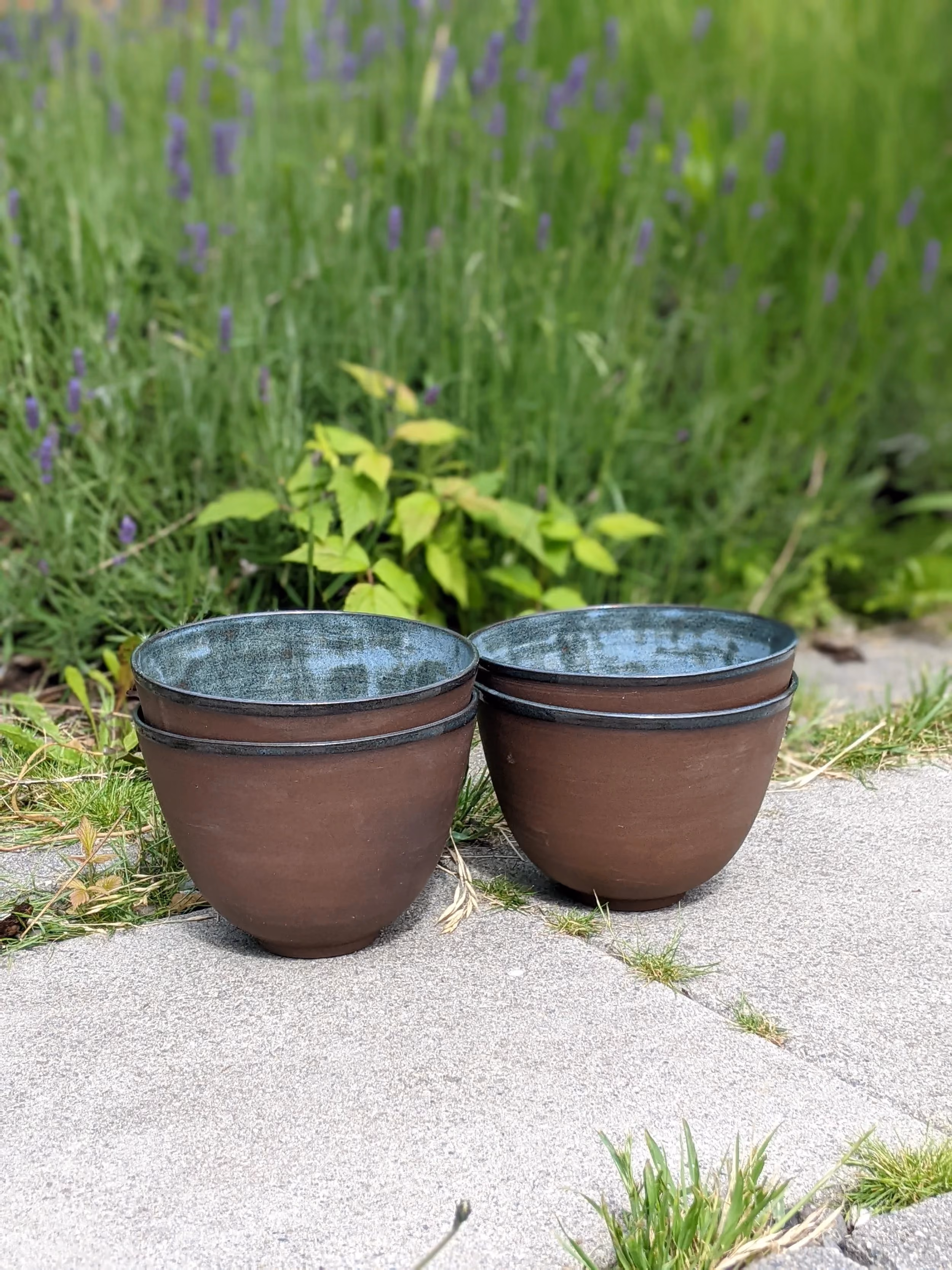 Two brown ceramic planter pots with dark glaze inside, placed on a concrete pathway with grass and garden plants in the background.