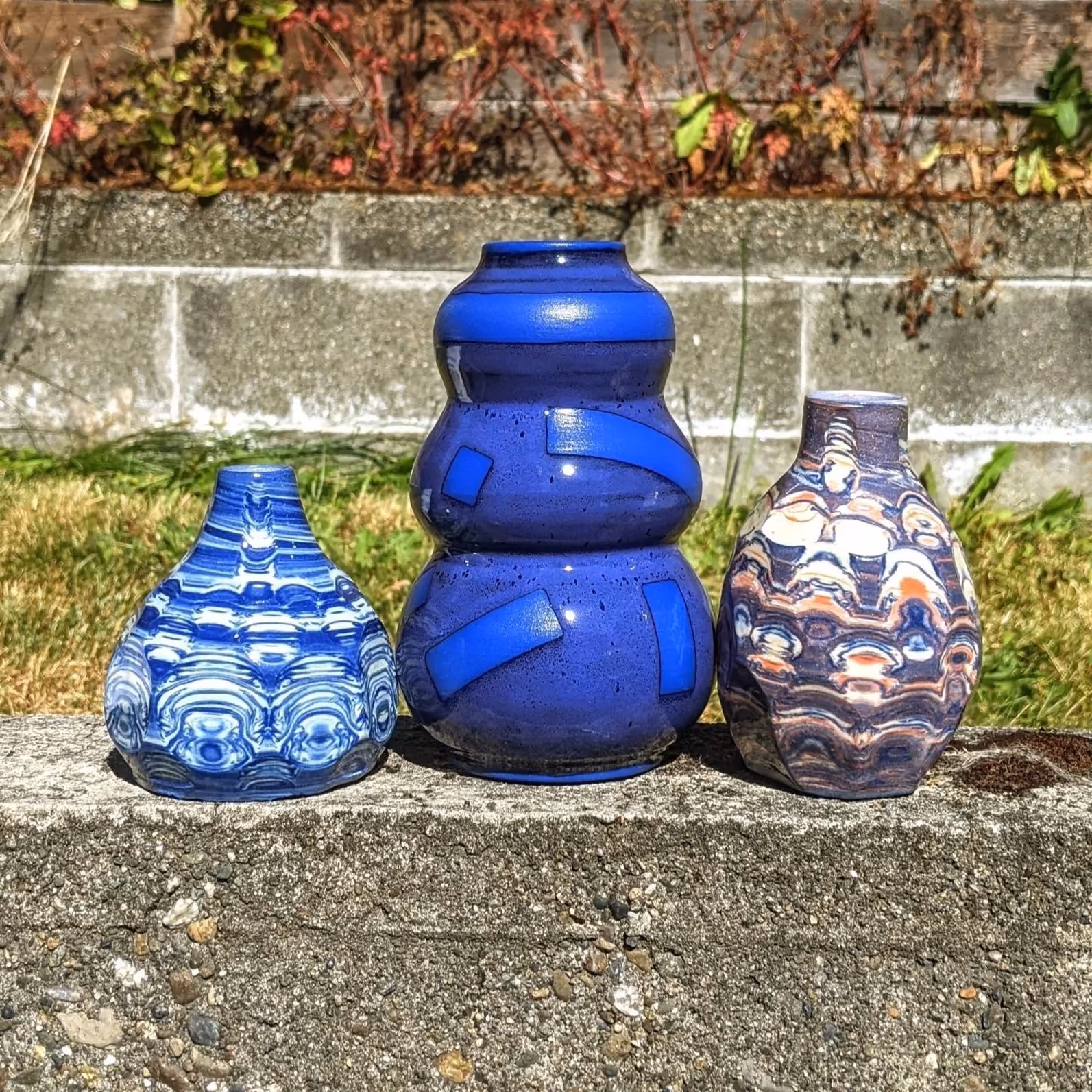 Three decorative glass vases on a concrete surface outdoors, with a brick wall and plants in the background. The central vase is large and blue, flanked by two smaller patterned vases.