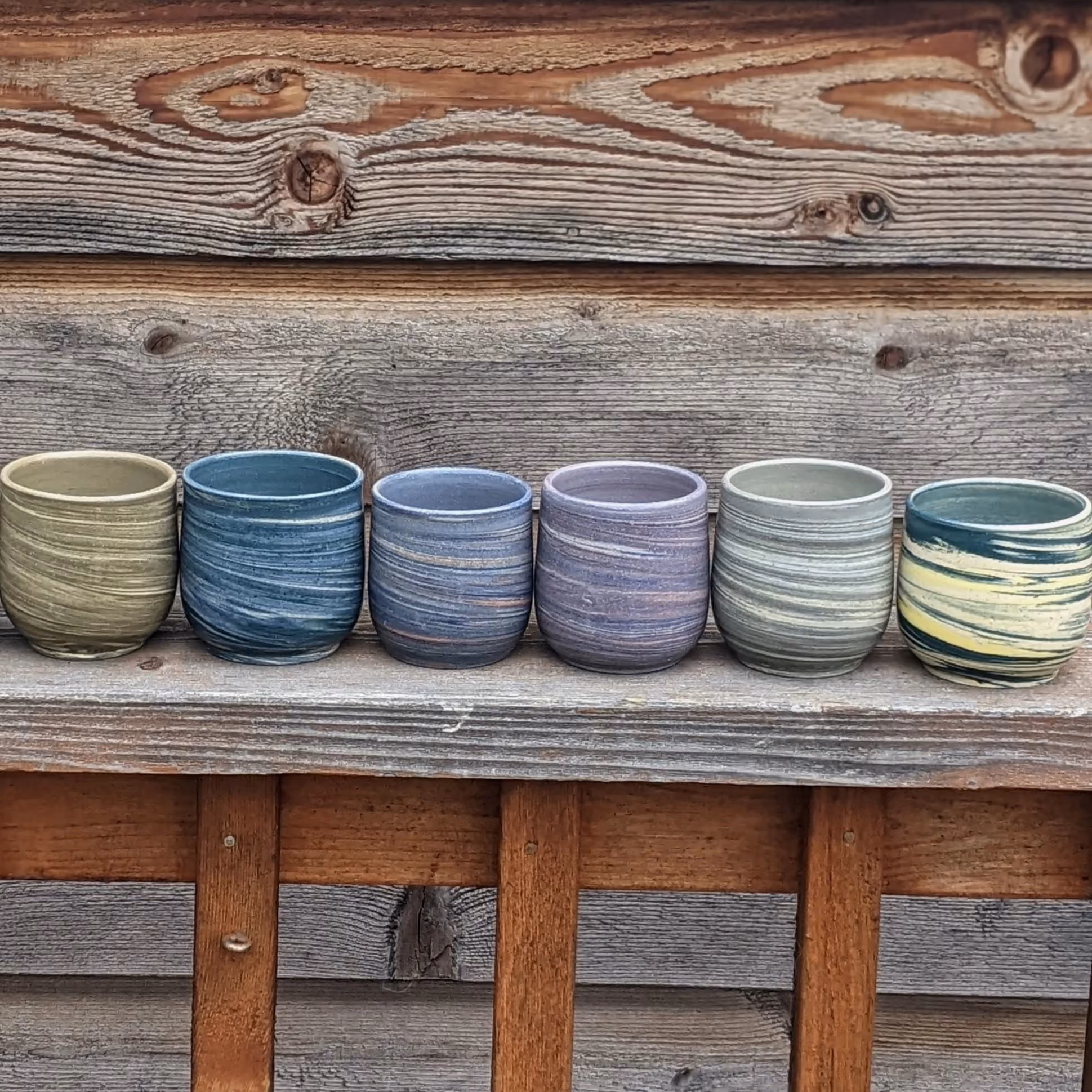 Six colorful, striped ceramic cups on a wooden shelf against a weathered wooden wall.