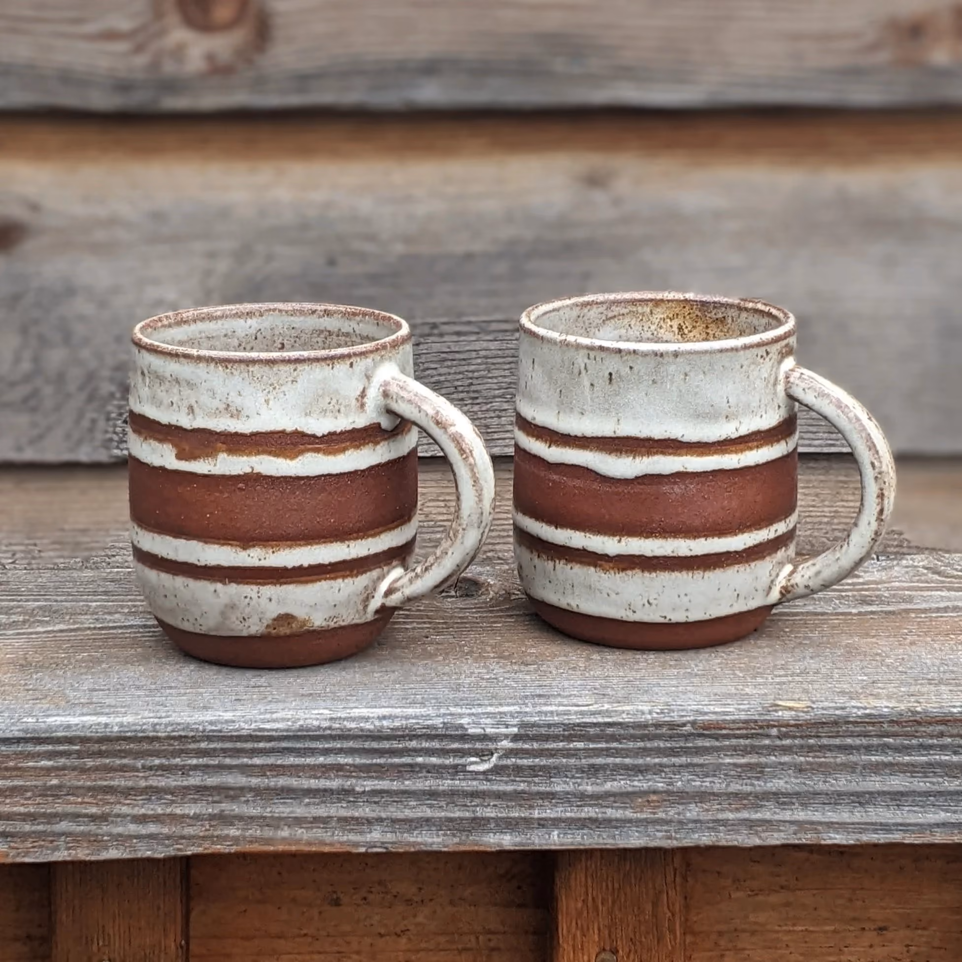 Two rustic ceramic coffee mugs with horizontal rust-colored and cream stripes, placed on a weathered wooden surface outdoors.