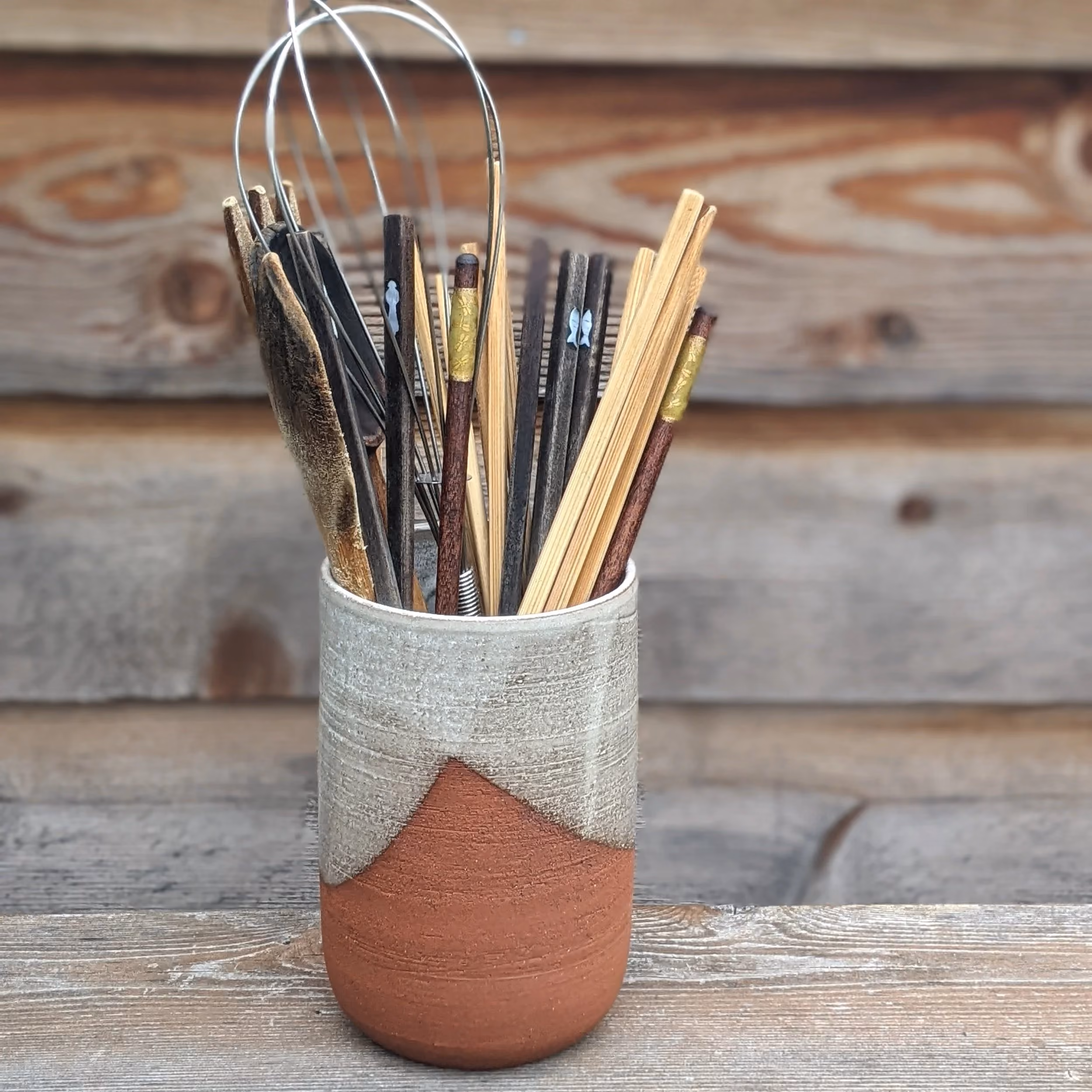 A ceramic mug filled with various wooden and metal kitchen utensils, such as chopsticks and whisks, placed on a wooden surface against a wooden wall background.