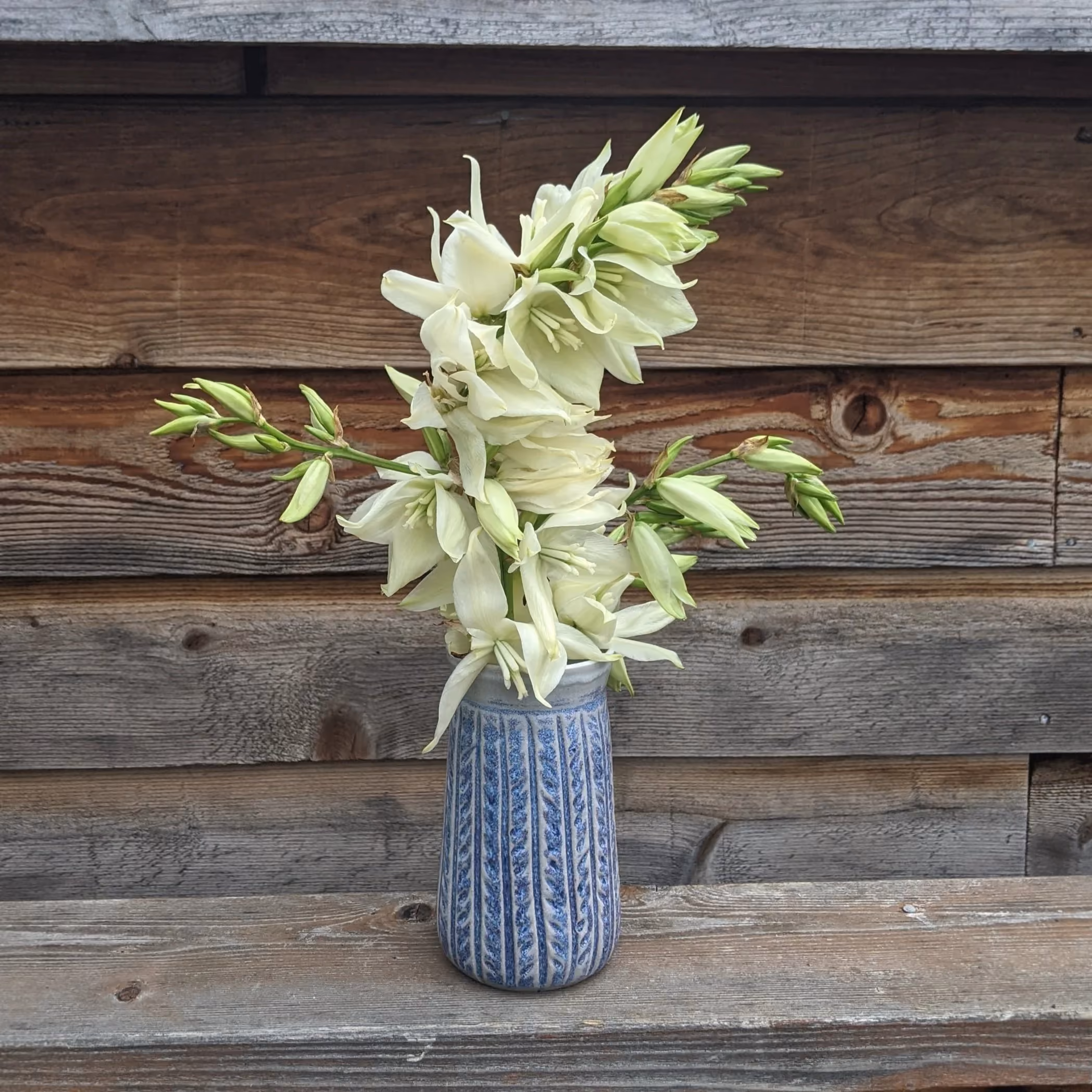 A blue and white patterned ceramic vase holding a bouquet of white flowers and buds, placed on a wooden surface against a wooden wall background.