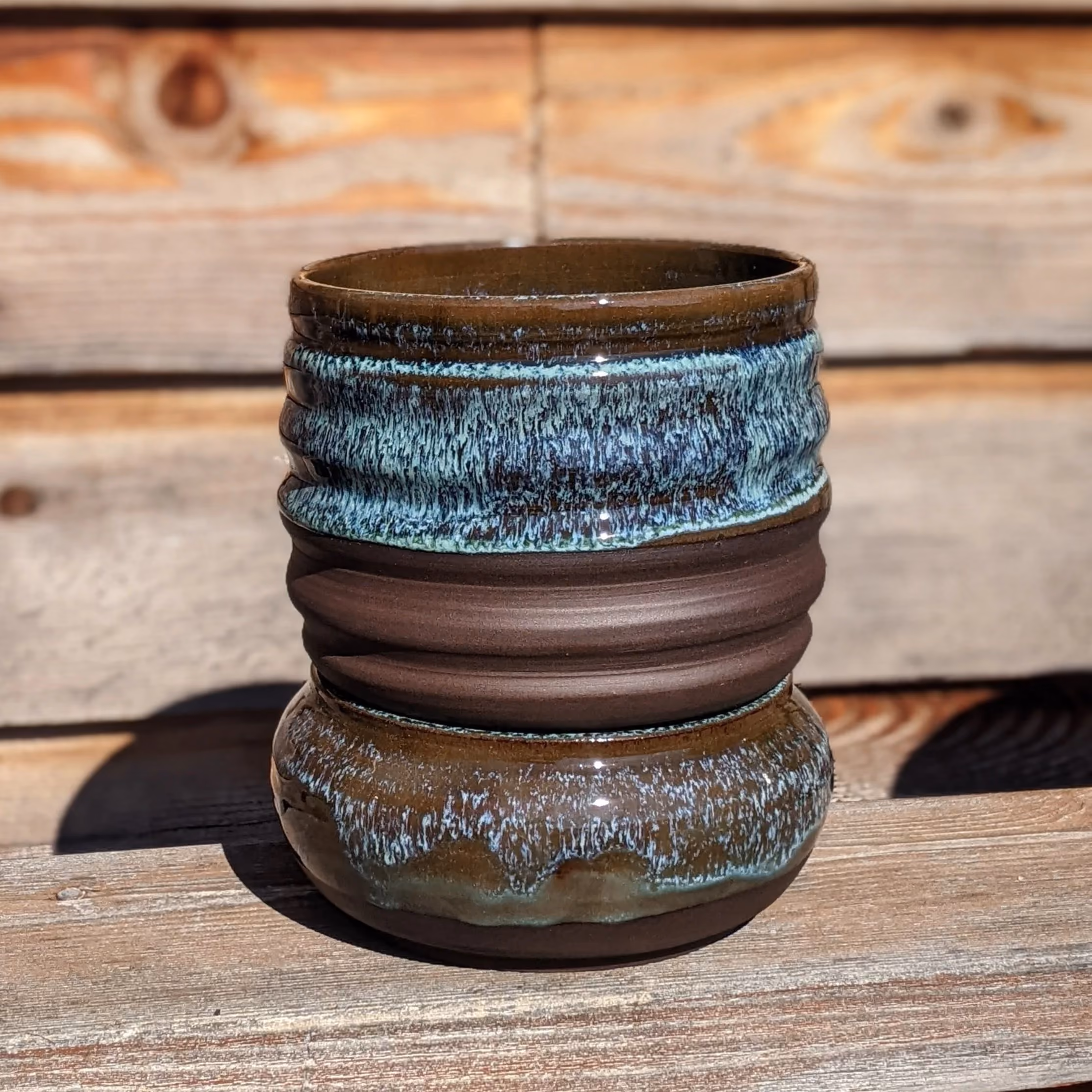 Ceramic pot with layered textures and blue glazed accents, placed on a wooden surface in sunlight.