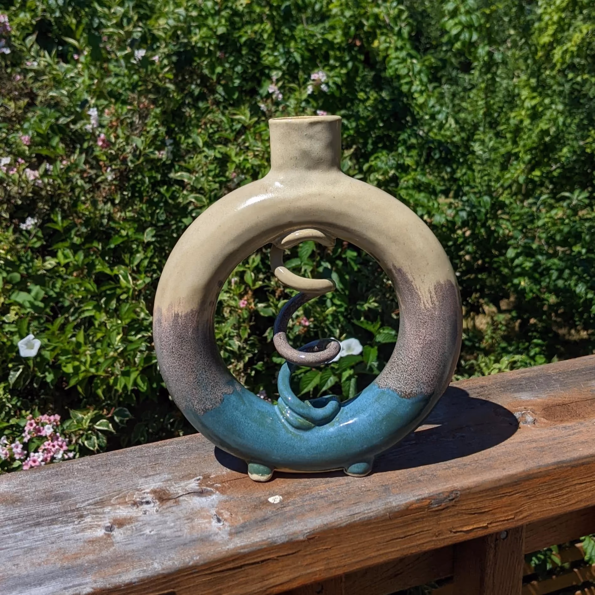 Ceramic sculpture with a circular shape, a small opening at the top, and a twisted spiral inside, placed on a wooden railing outdoors with green foliage in the background.