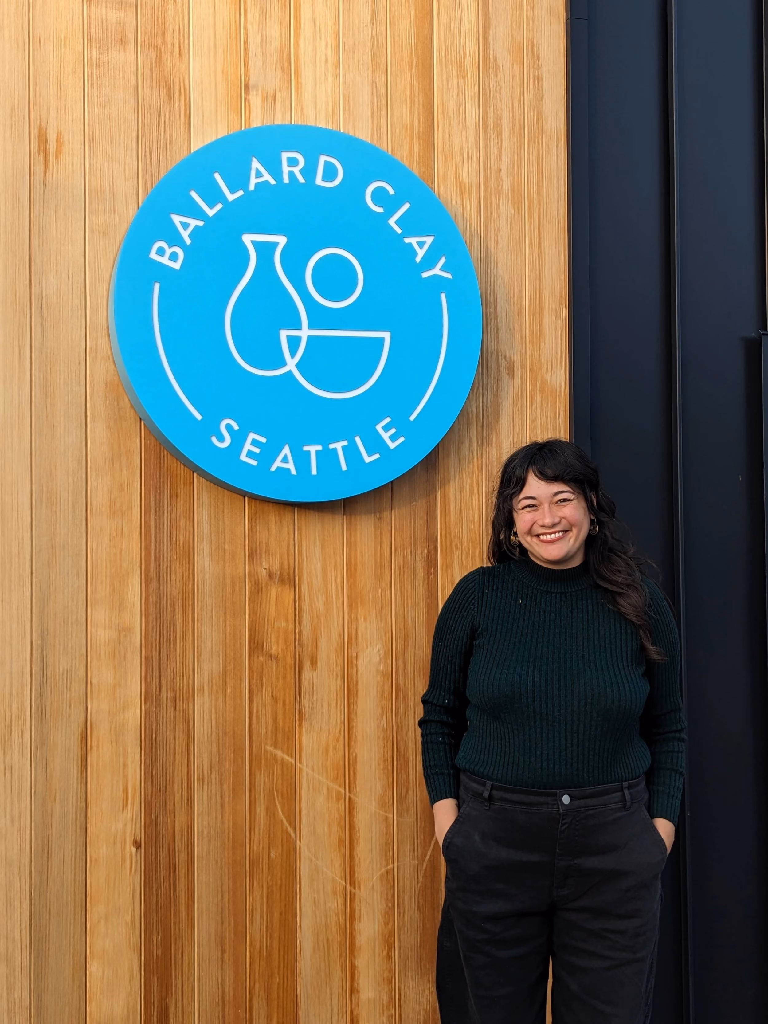 A woman standing in front of a wooden wall with a blue circular sign that reads “Ballard Clay Seattle.” The woman is smiling, wearing a dark green ribbed sweater and black pants.