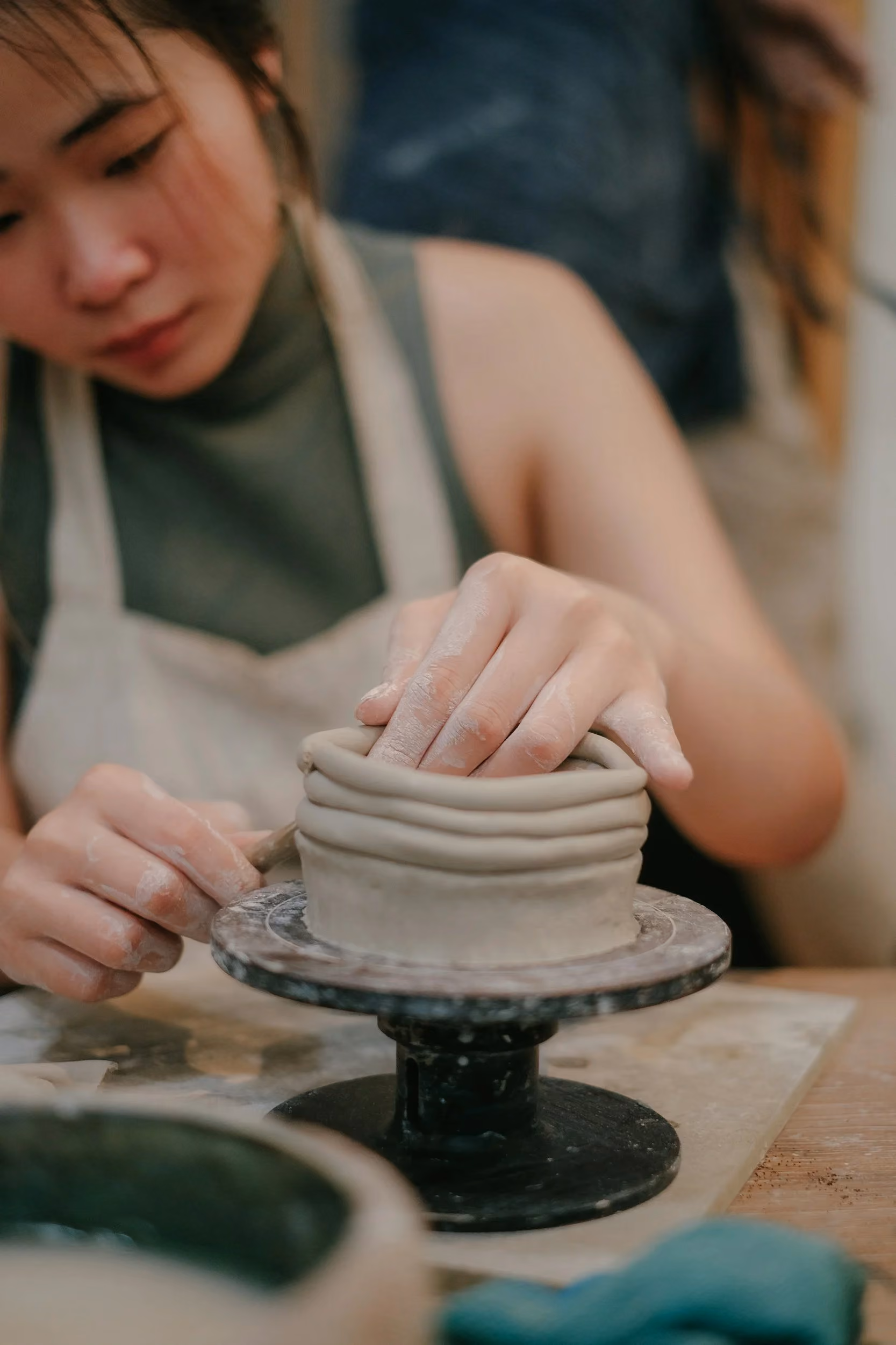 A student in a canvas apron shaping a small coil-built clay vessel on a turntable.
