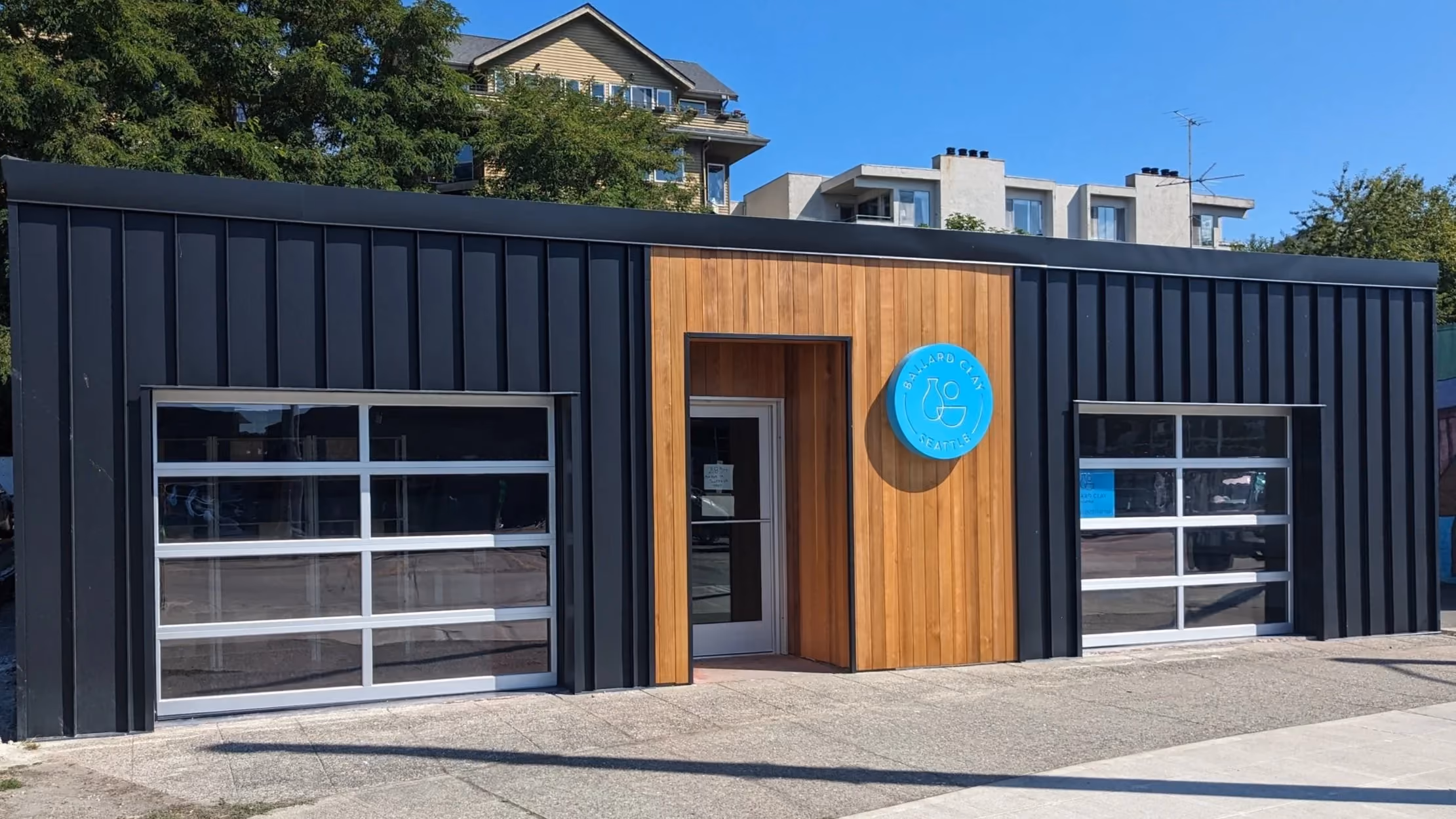 The Ballard Clay studio from the street — a black corrugated building with a cedar entrance and the round blue studio badge beside the door.