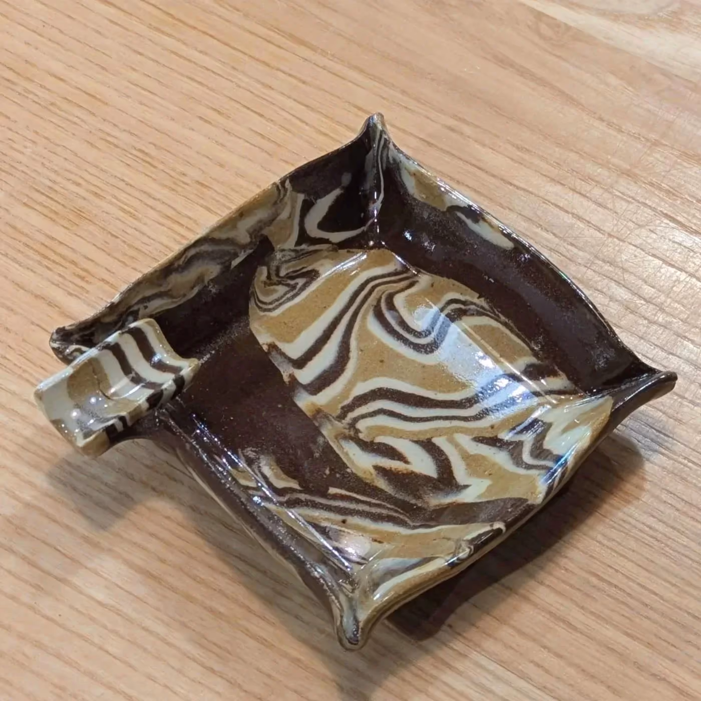 A square hand-formed dish with pinched corners, its surface marbled in swirling bands of dark brown, cream and tan clay.