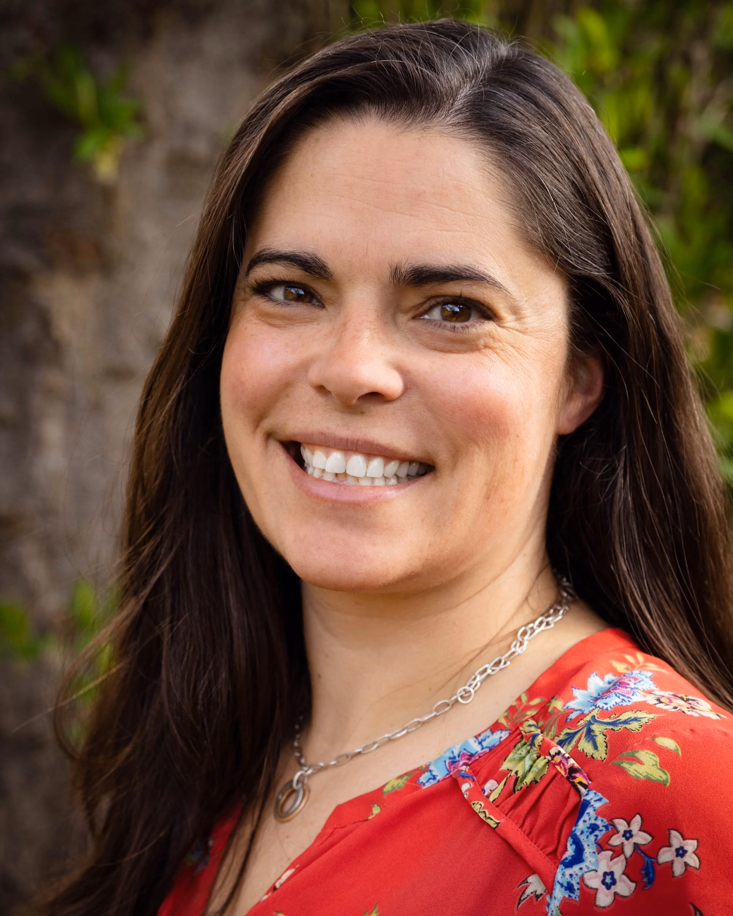 Rebecca Sorensen photographed outdoors in front of a stone wall and greenery, smiling, wearing a red floral top.