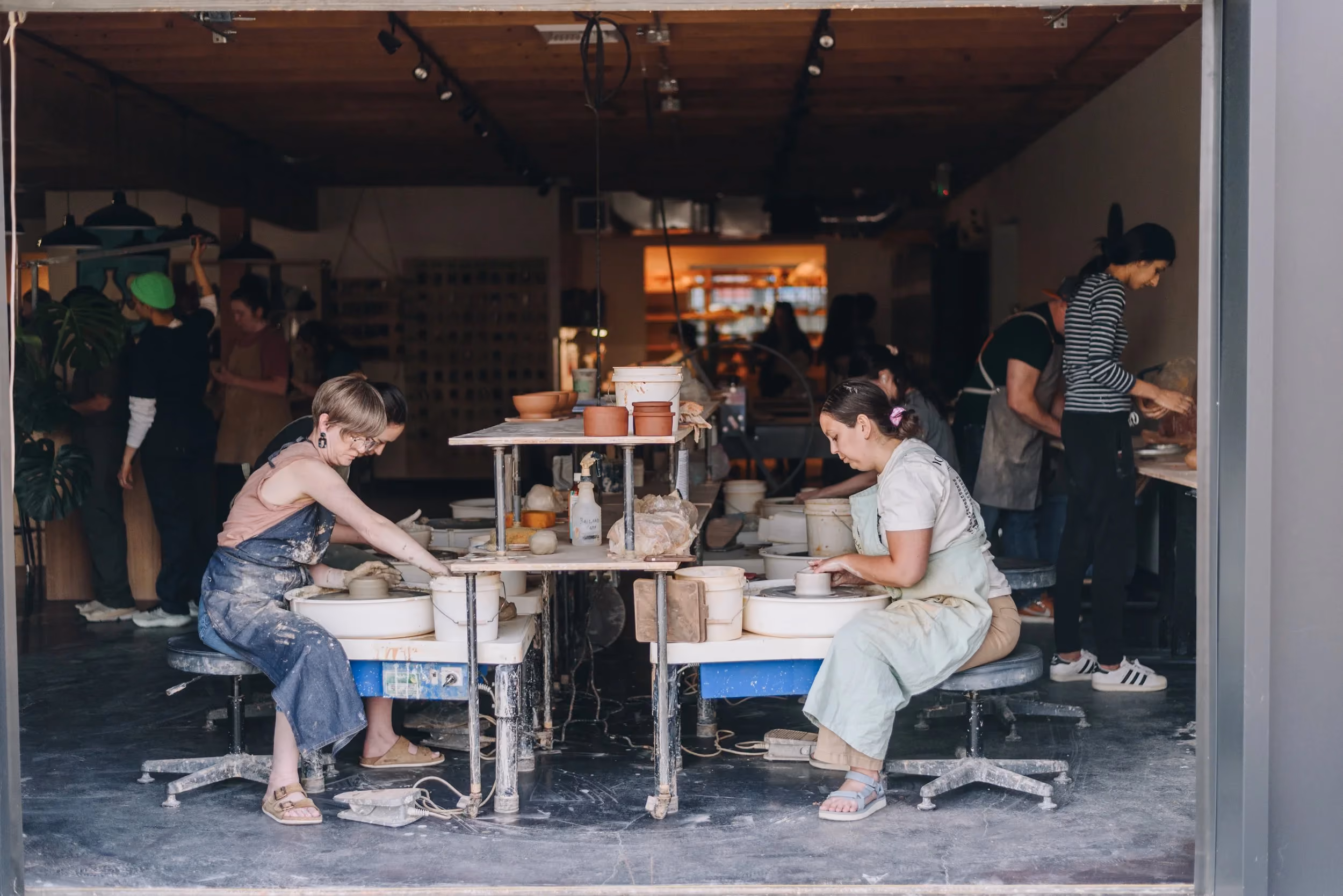 Two potters working at wheels inside the studio, seen through the open garage door from the street.