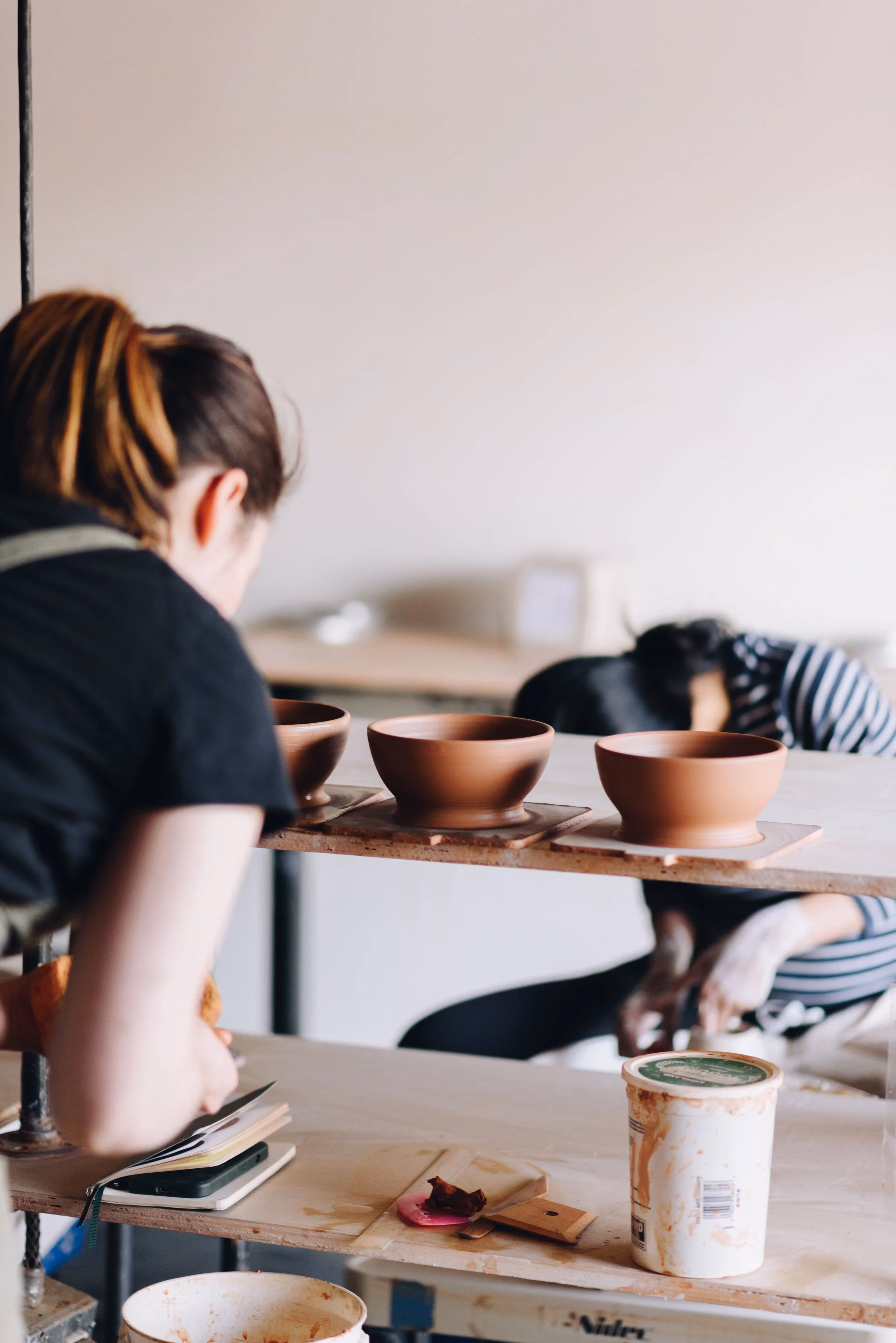 People working on pottery projects at a ceramics studio with clay bowls and tools on tables.